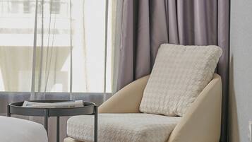 Habitación de esquina con cama, una silla con cojín, una mesita y una ventana con cortinas.