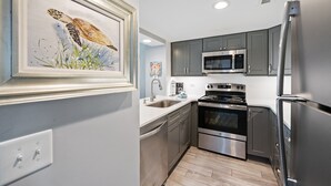 A modern kitchen with stainless steel appliances, grey cabinets, and a framed seascape painting. - Recently Renovated Oceanfront Condo, Private Resort +Free Attraction Tickets! (Myrtle Beach)