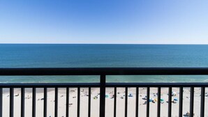 Uitzicht op een strand vanaf een balkon met een hekwerk, helderblauwe lucht en een rustige zee.