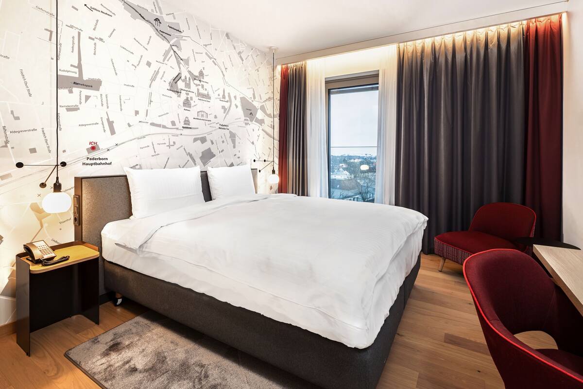 A hotel room with a large bed, a desk, a chair, a map wall, and a view of the city.