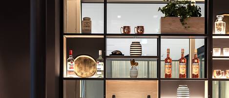 A bar area with shelves displaying bottles and glasses, a wooden counter, and a seating area with chairs.