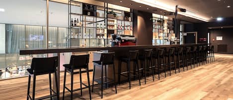 Zona de bar moderna con una barra larga, taburetes negros y una variedad de botellas y vasos en estantes.