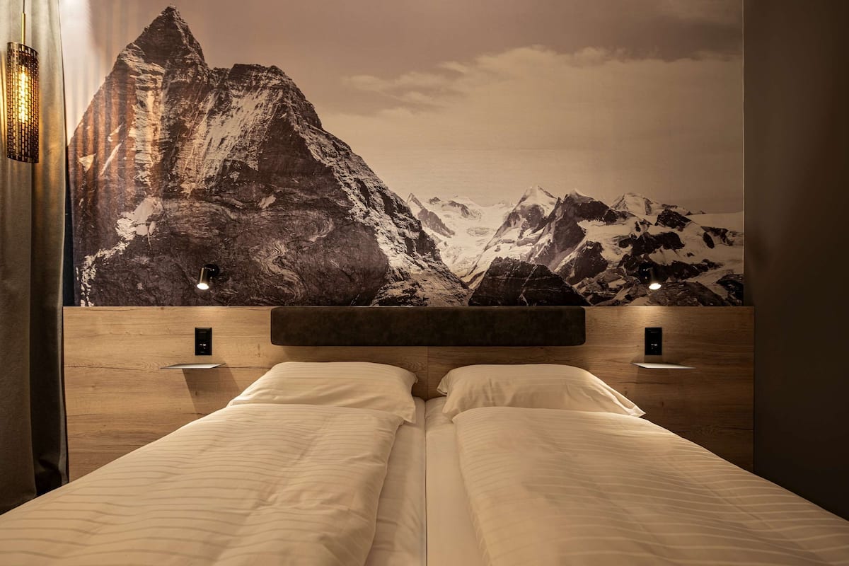 Ein Hotelzimmer mit einer großen Wandmalerei einer Bergkette, einem Holzkopfende, zwei Nachttischen mit Lampen und einem Bett mit weißen Leinen.