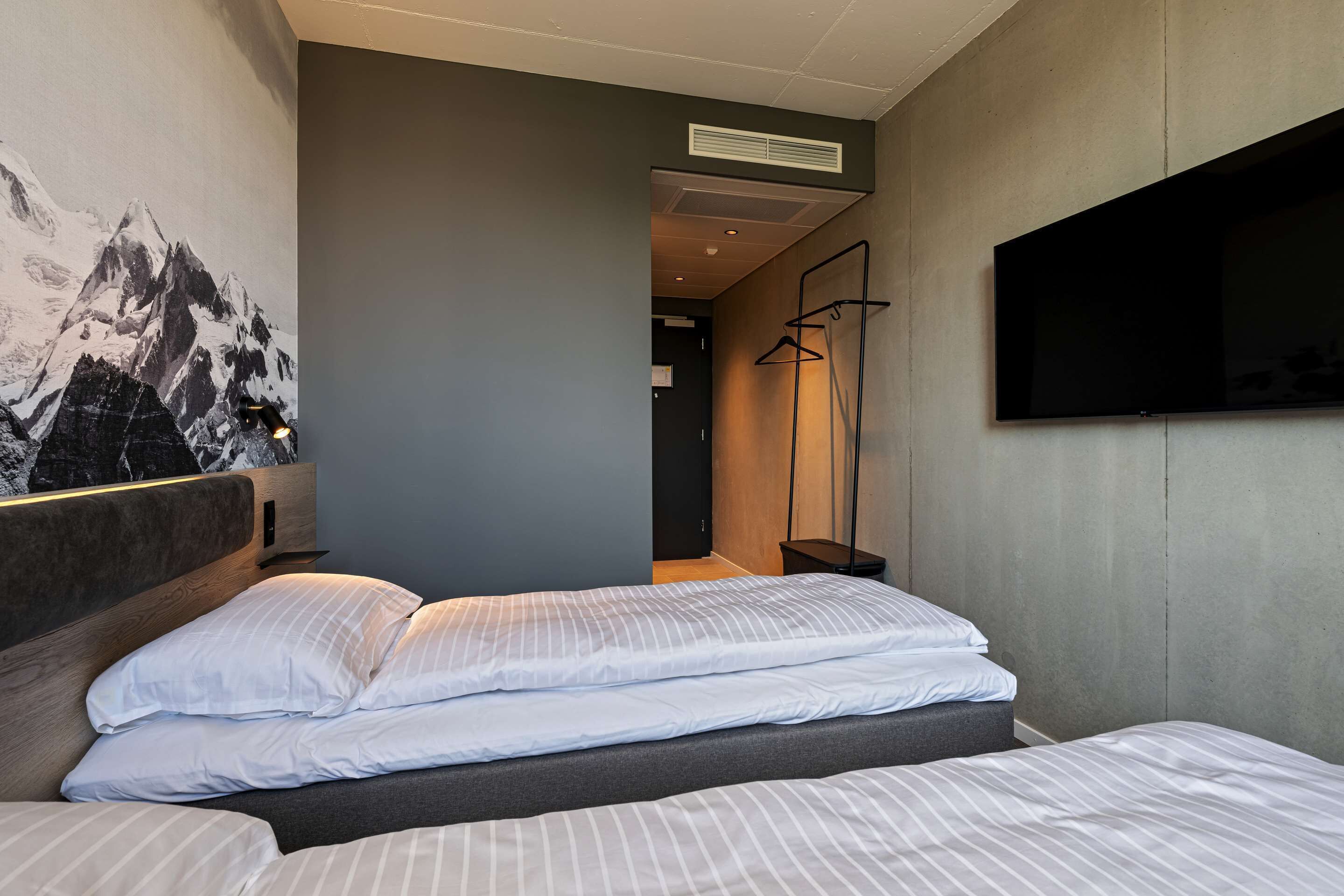 A hotel room with a bed, a mounted TV, and a painting of mountains on the wall.