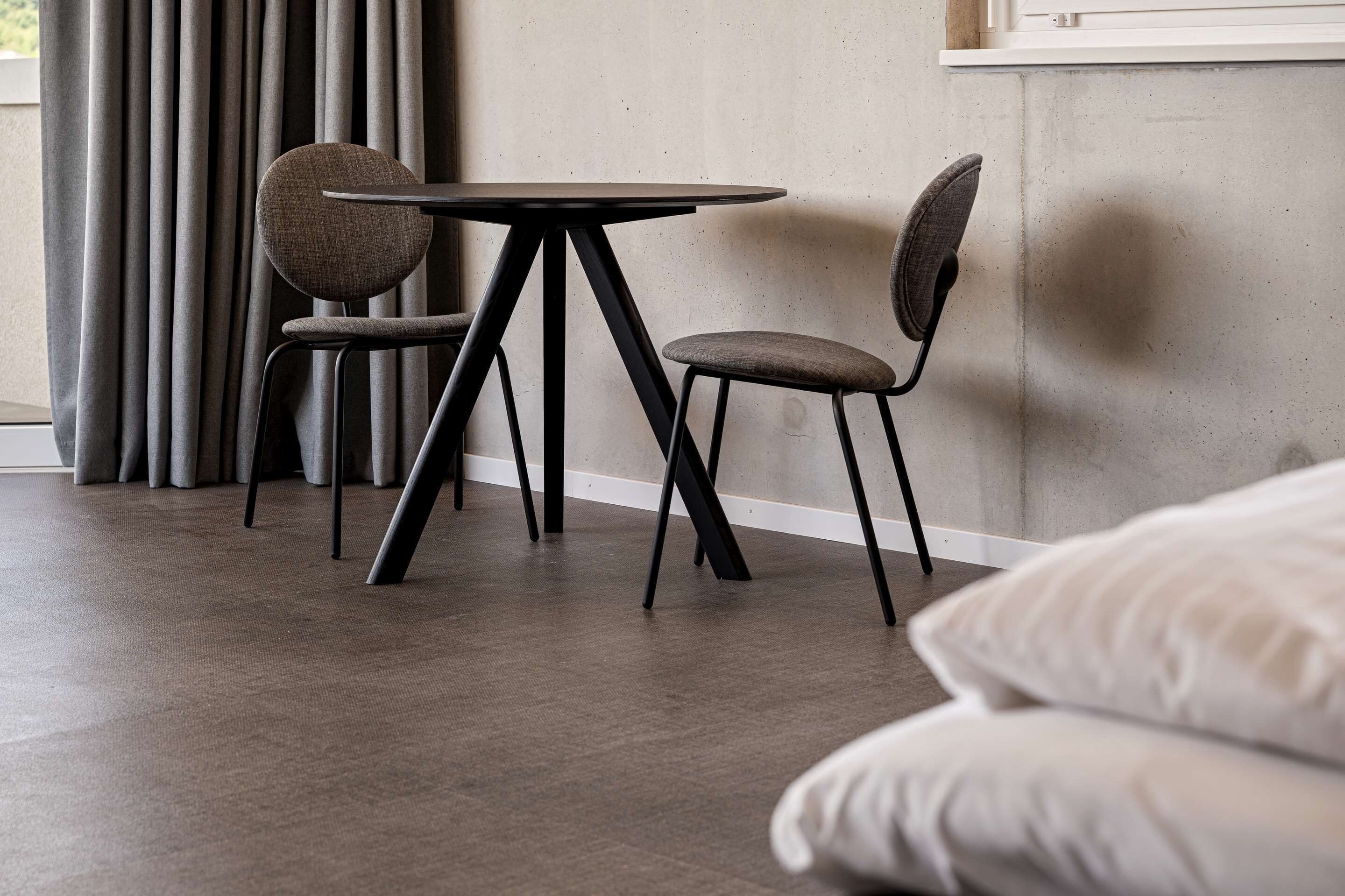 Una habitación moderna con una mesa redonda y dos sillas, un ventanal con cortinas y una pared de hormigón.