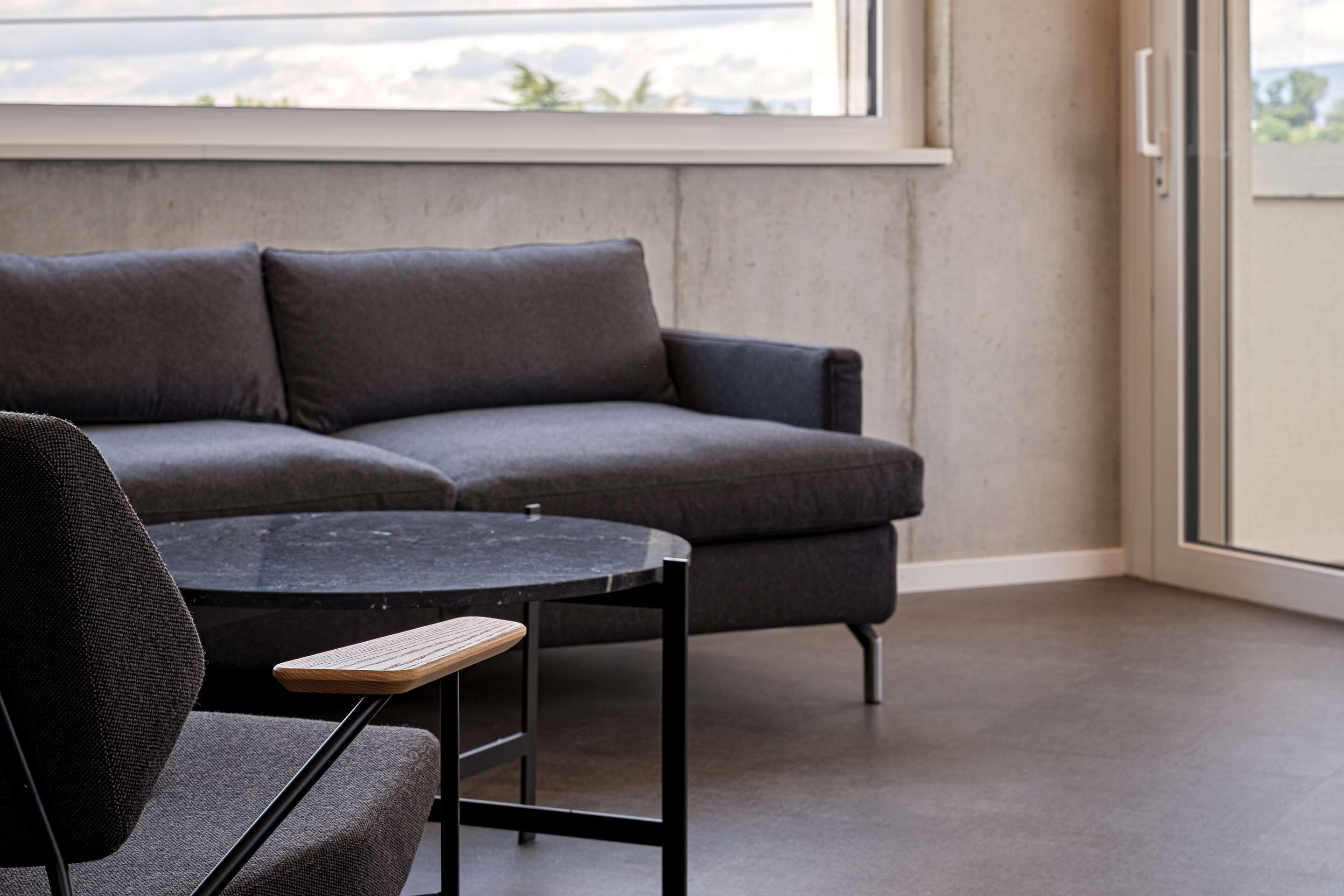 Ein modernes Wohnzimmer mit einem grauen Sofa, einem runden schwarzen Tisch und einem Holzstuhl.