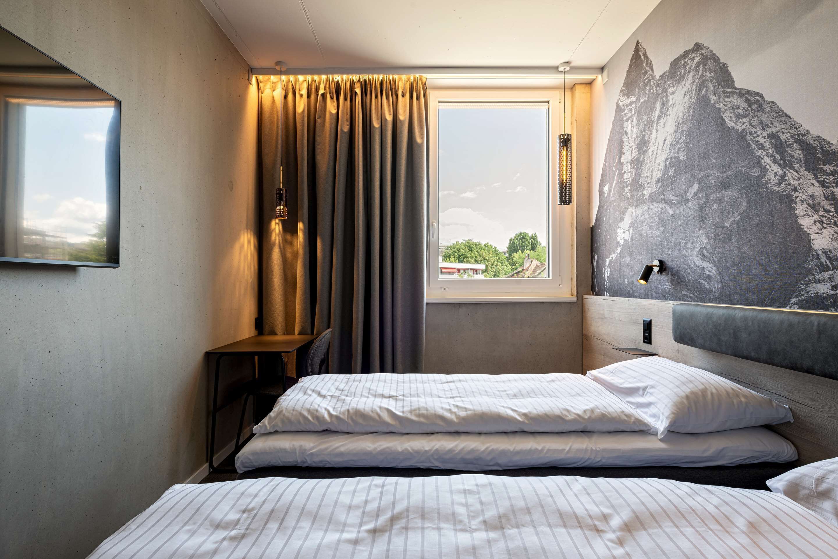 A hotel room with a bed, a window with curtains, a wall-mounted lamp, and a large mountain mural.