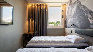 A hotel room with a bed, a window with curtains, a wall-mounted lamp, and a large mountain mural.