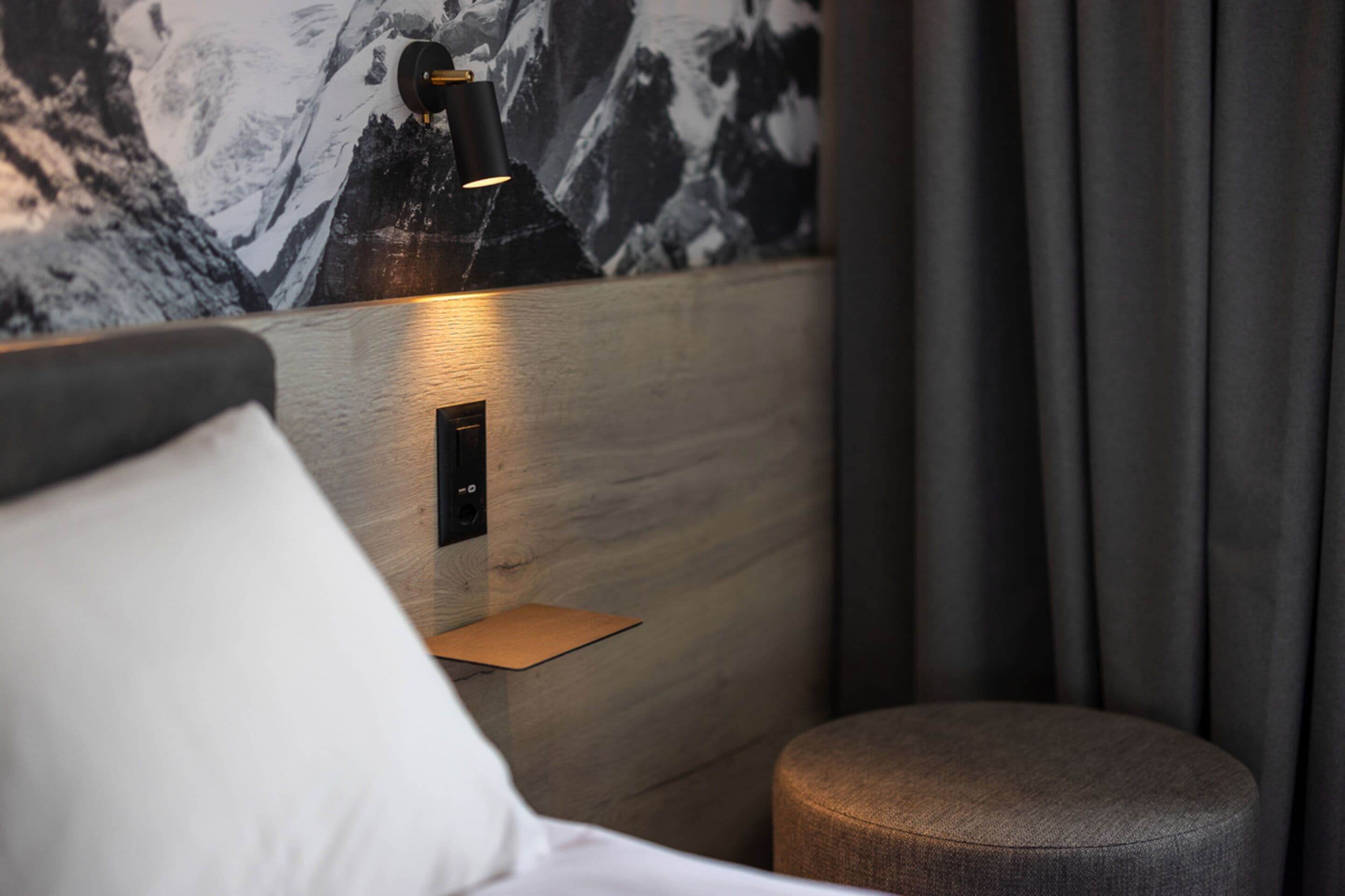 Et moderne hotelværelse med en seng, et natbord, en vægmonteret lampe og en væg med et bjergmotiv.