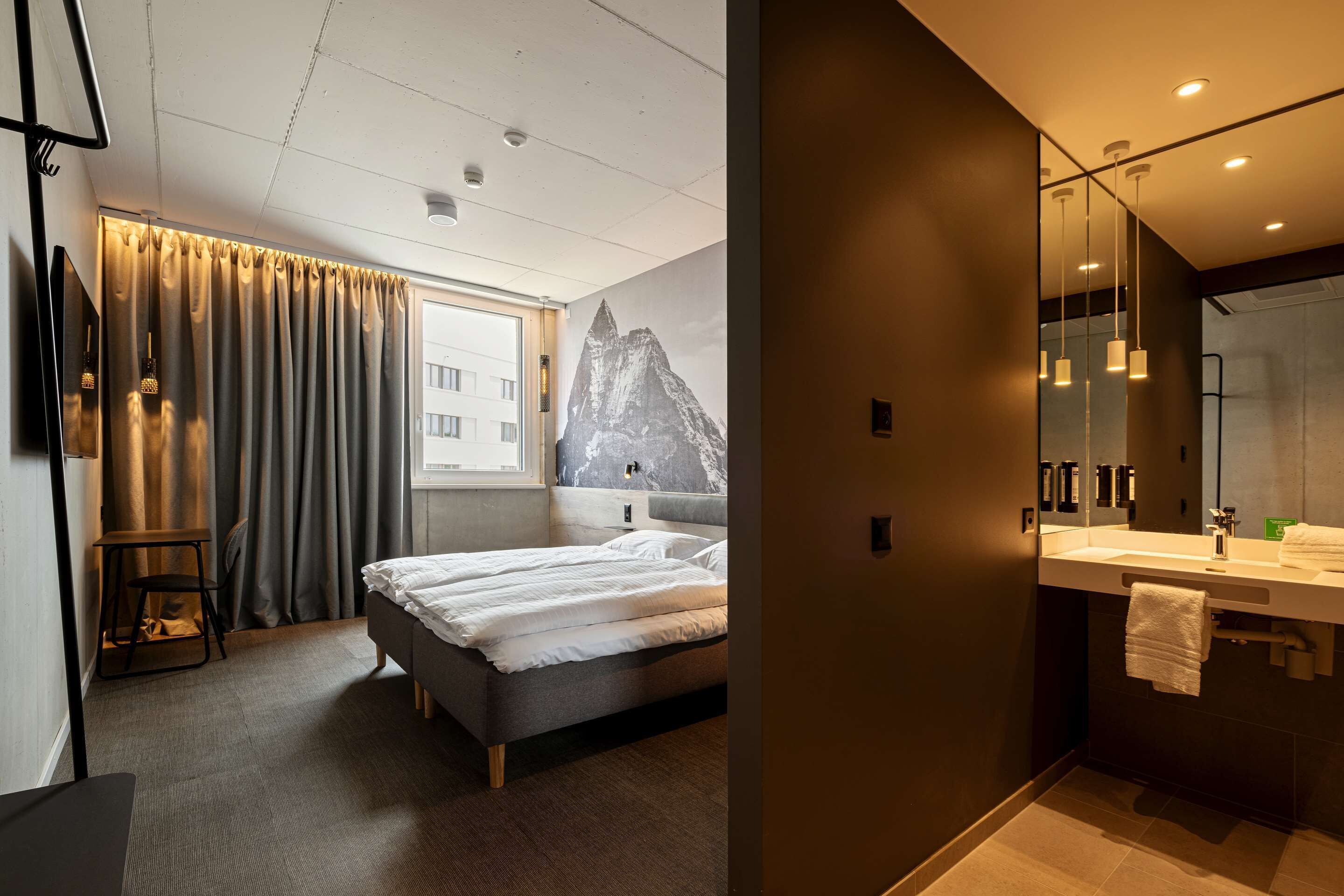 Une chambre d’hôtel moderne comprenant un lit, une grande fenêtre avec des rideaux, un tableau représentant une montagne, une salle de bain avec un miroir, un lavabo et un porte-serviettes.