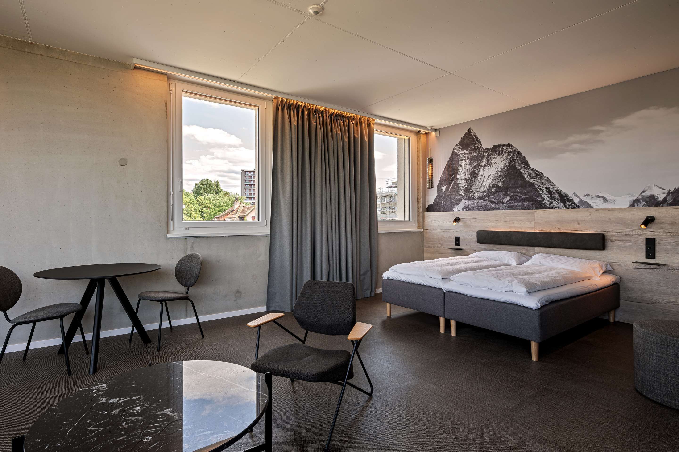 A modern hotel room with a bed, two chairs, a small table, and a large window with curtains.