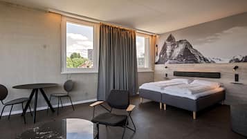 A modern hotel room with a bed, two chairs, a small table, and a large window with curtains.