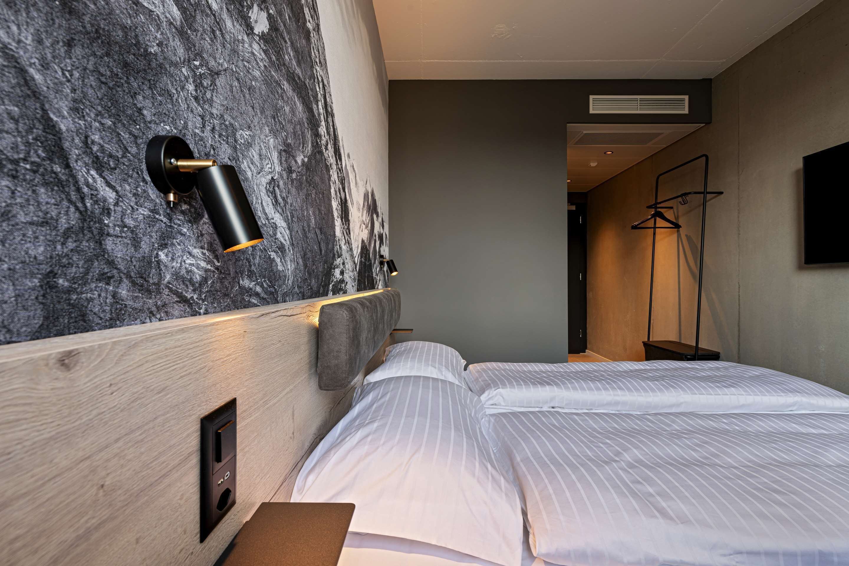 A modern hotel room with a large bed, a headboard with a marble-like texture, a wall-mounted light, and a coat rack.