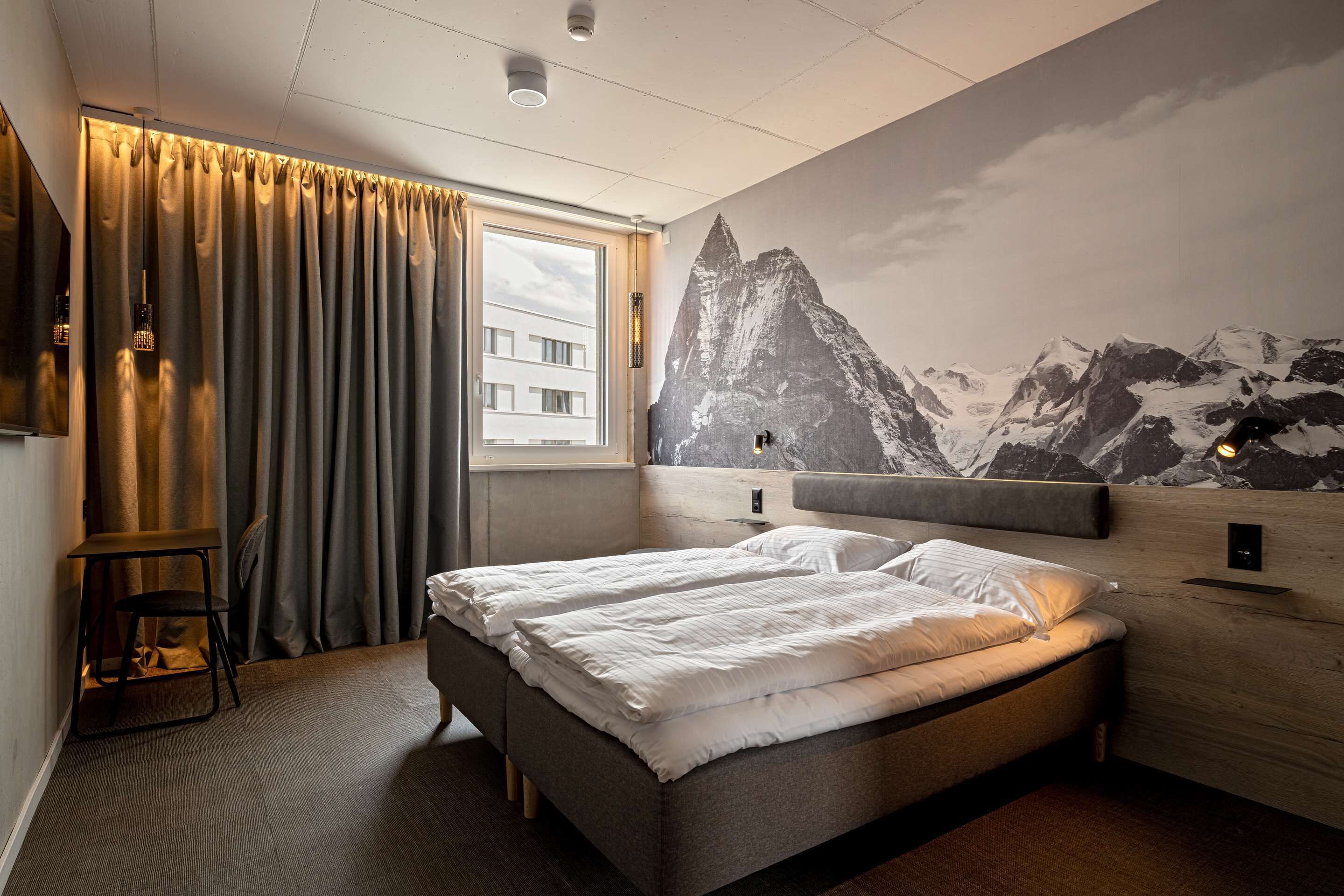 A hotel room with a bed, a desk, a chair, a window with curtains, and a wall mural of mountains.