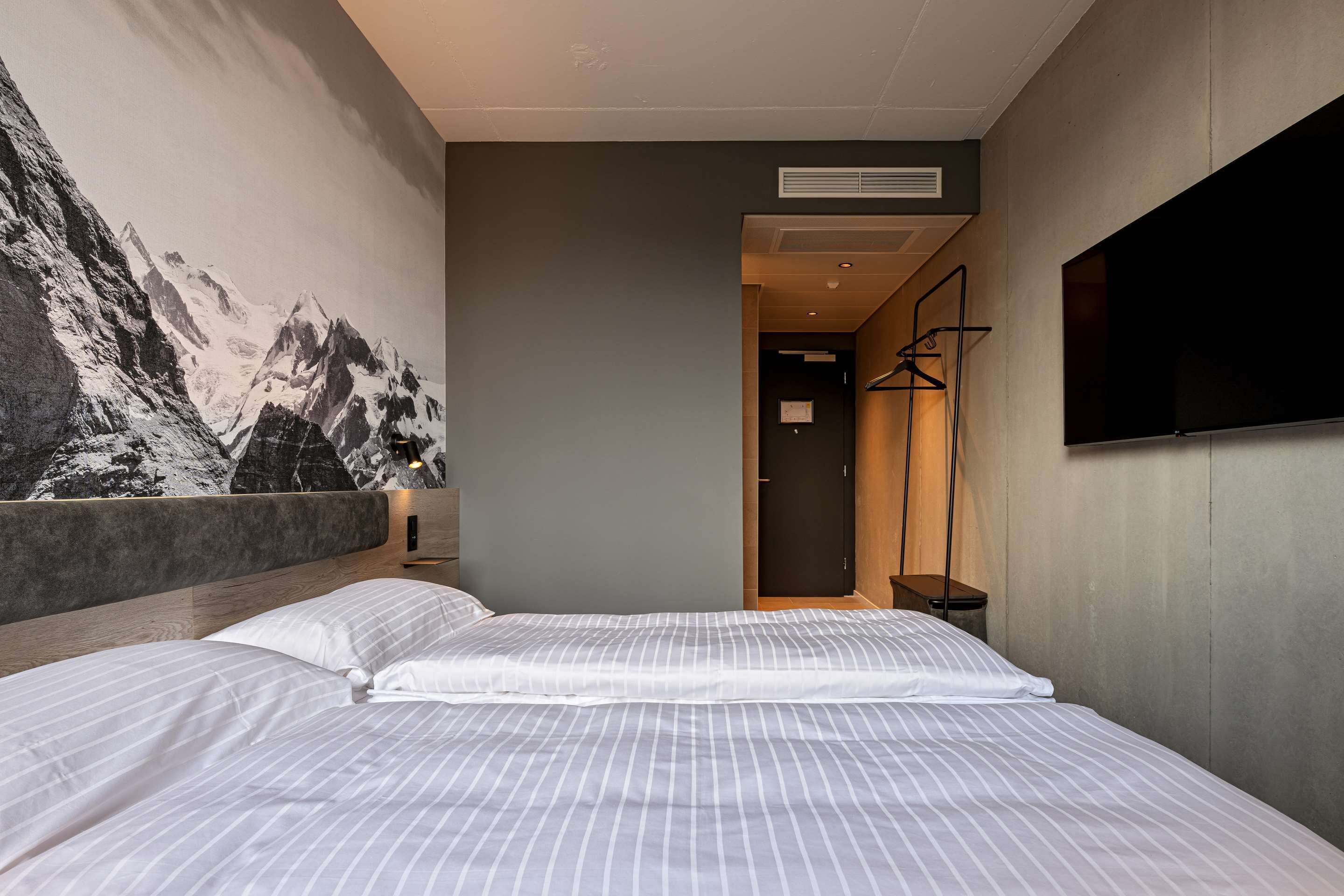 A modern hotel room with a large bed, a mounted TV, and a mountain mural on the wall.