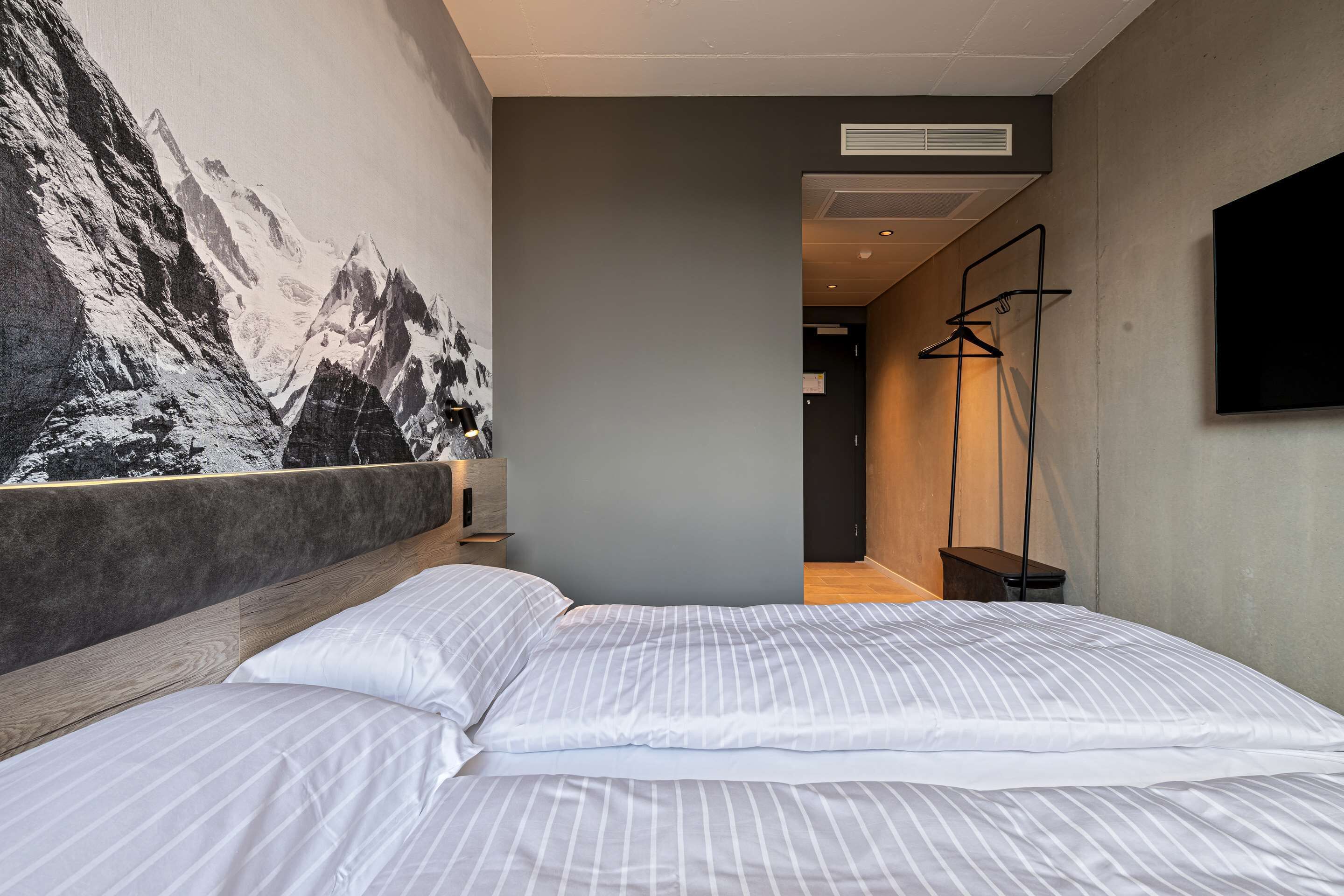 A modern hotel room with a large bed, a wall-mounted TV, and a mountain painting.