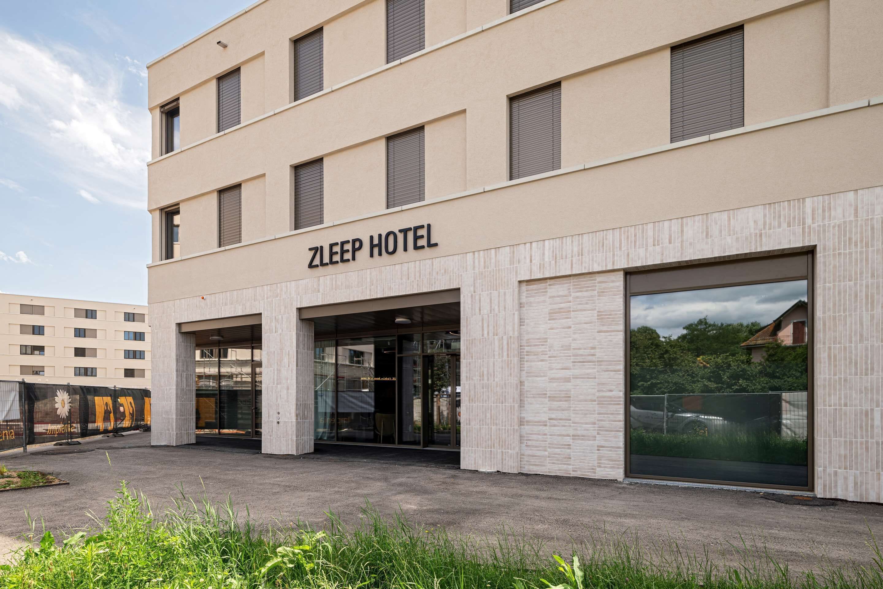 Un moderno hotel Zleep con una fachada limpia, un amplio acceso de vidrio y un área de estacionamiento al frente.
