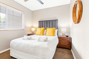 A hotel room with a large bed, two bedside tables, a ceiling fan, a window with blinds, and a wooden wall decor. - Lagoon Beach 29A (Cape Town)