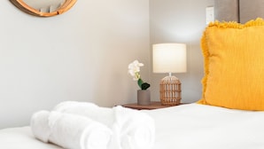 A neatly made bed with rolled white towels, a bedside lamp, a small potted plant, and a yellow pillow. - Lagoon Beach 29A (Cape Town)