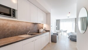 Une cuisine moderne équipée d’un four à micro-ondes encastré, d’un évier et d’un plan de travail. On y trouve également un coin repas avec une table et des chaises, ainsi qu’un lit avec une tête de lit grise en arrière-plan.
