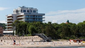Un hôtel à plusieurs étages doté de balcons, une plage avec un escalier, et une colline boisée en arrière-plan.
