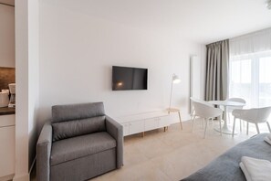 A modern living room with a sofa, a flat-screen TV mounted on the wall, a dining area with a table and chairs, and a large window with curtains.