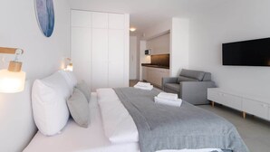Una habitación de hotel moderna con una cama, un sofá gris, un televisor de pantalla plana y una kitchenette al fondo.