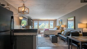 A modern kitchen with dark cabinets and a granite countertop, a dining area with a wooden table and chairs, a cozy bedroom with a bed and a nightstand, and large windows offering a view of trees. - The Serenity Studio | AC, Pool, Hot Tub, Golf & Ski (Durango)