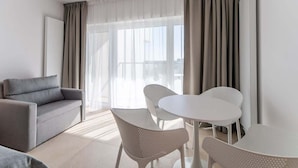 A modern hotel room with a grey sofa, a small round table, two white chairs, and a large window with sheer curtains.