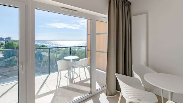 A balcony with a view of a body of water and greenery, furnished with white chairs and a table.