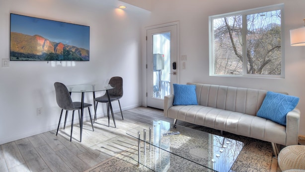 Una sala moderna con una mesa de vidrio, dos sillas, un sofá con cojines azules, un cuadro en la pared y un ventanal con vistas a árboles.