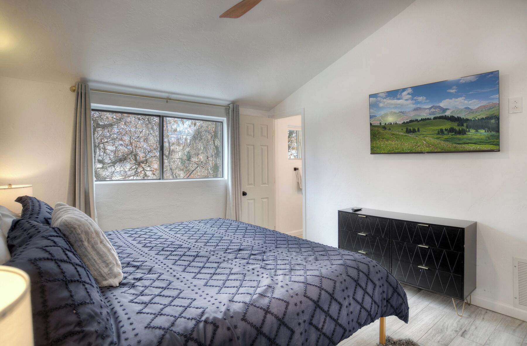 Ein Schlafzimmer mit Bett, Fenster, Tür, Kommode und Landschaftsbild an der Wand.