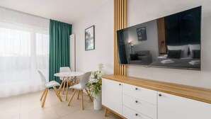 Una sala de estar moderna con un televisor de pantalla plana montado en la pared, un comedor con sillas blancas y una mesa redonda, y un amplio ventanal con cortinas translúcidas.