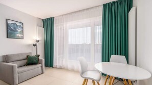 Una sala de estar moderna con un sofá gris, sillas blancas, una mesa redonda y cortinas verdes.