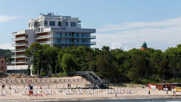 Un hotel moderno de varios pisos con balcones, situado cerca de una playa con una escalera que baja hasta la orilla.