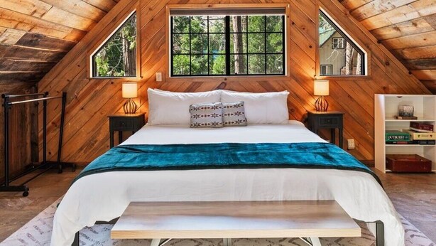 Un dormitorio con techo de madera, una cama con manta azul, dos mesitas de noche con lámparas y una ventana con vistas a árboles.