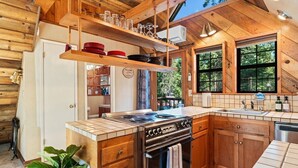 Cocina de cabaña de madera con estufa central, armarios de madera y ventana con vistas a la vegetación.