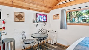 A cozy room with a wooden ceiling, a TV mounted on the wall, a welcome sign, a window with curtains, and a small table with chairs.