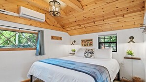 A cabin-style bedroom with a wooden ceiling, a bed with a blue and white bedspread, a 'Welcome to the Cabin' sign, and a small plant on a shelf.