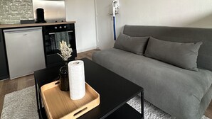 Un salon moderne avec un canapé gris, une table basse noire surmontée d’un plateau en bois, un petit vase contenant des fleurs séchées et un miroir rond accroché au mur.