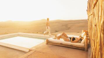 Un espace piscine avec une terrasse en bois, une chaise longue et une personne qui passe.