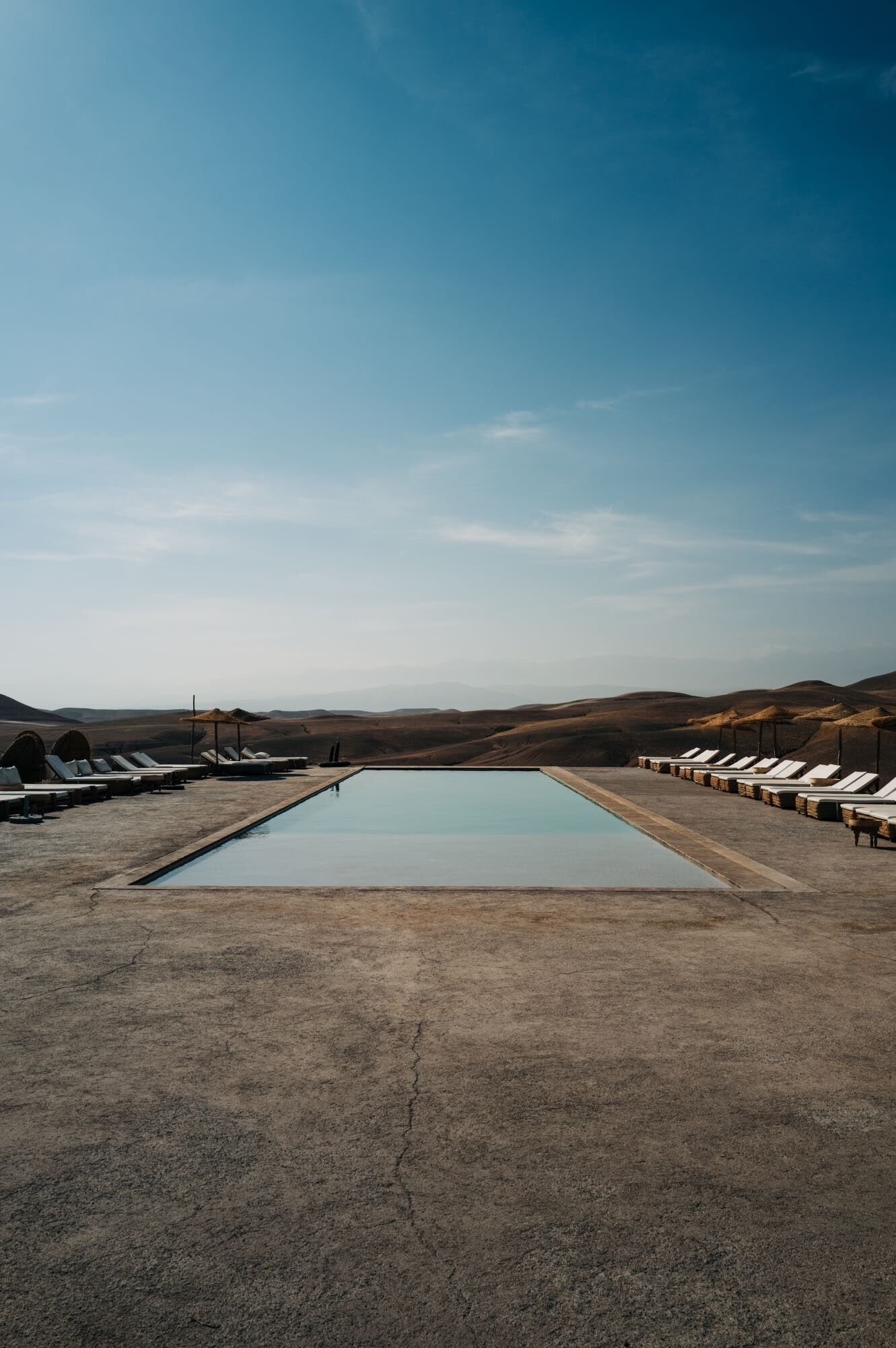 A rectangular pool with lounge chairs on both sides, set against a backdrop of rolling hills under a clear sky.