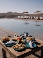 A dining table with various dishes and a teapot by a poolside with lounge chairs and umbrellas in the background.
