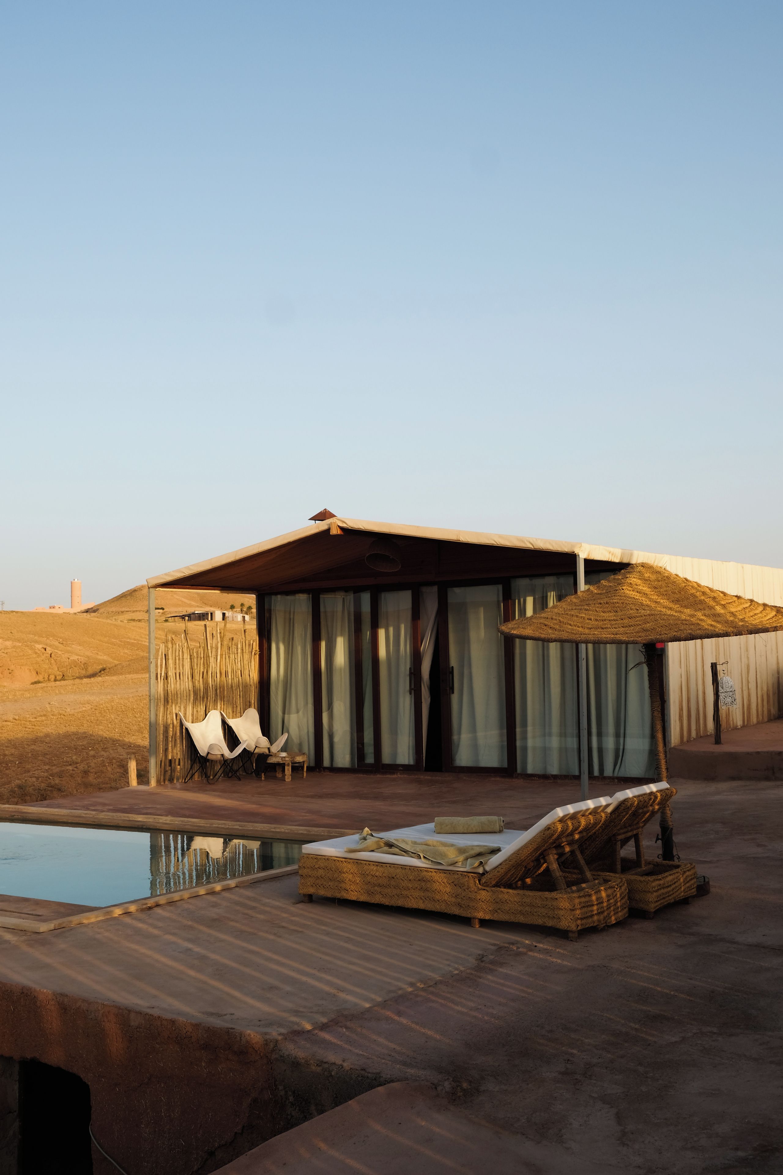 A poolside area with a lounge chair, a small table, and a covered seating area with curtains.
