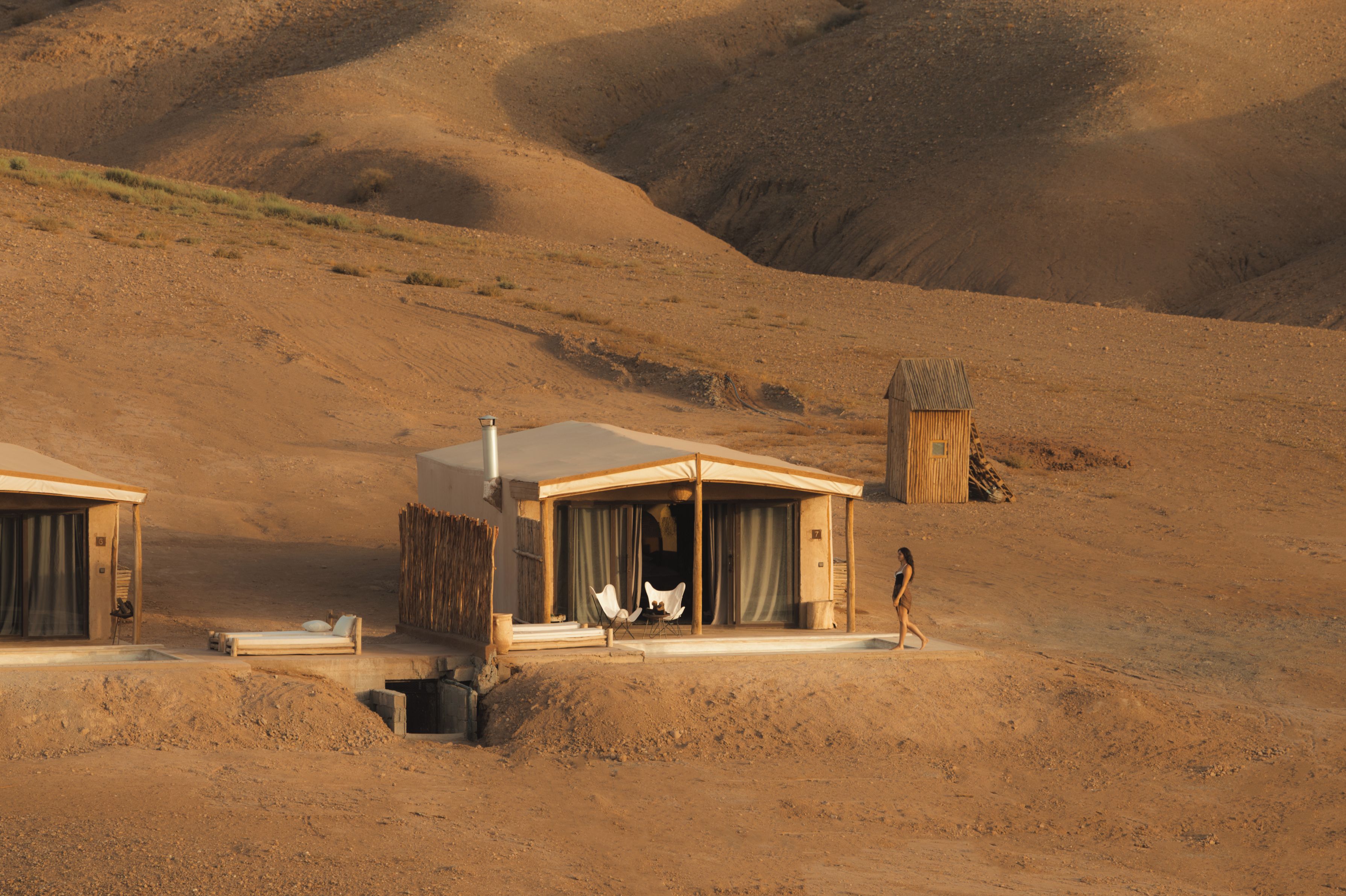 Paisaje desértico con dos cabañas de madera, una con un porche cubierto y una pequeña mesa, y la otra con una chimenea.