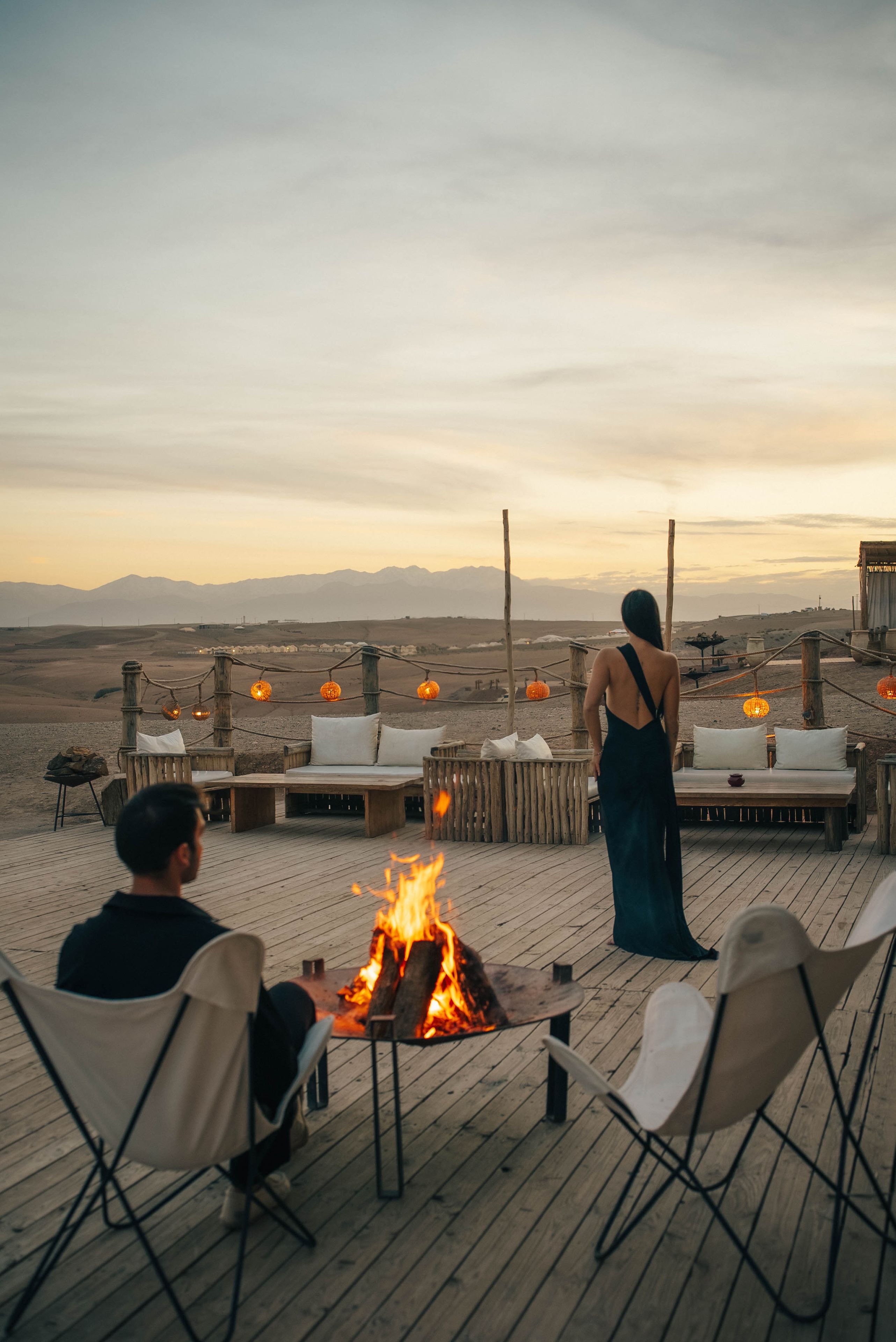 Una terraza de madera con mobiliario exterior y un pozo de fuego, con vistas a un paisaje desértico y montañas al fondo.