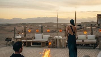 Una terraza de madera con mobiliario exterior y un pozo de fuego, con vistas a un paisaje desértico y montañas al fondo.