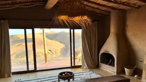 A room with a large window overlooking a desert landscape, a fireplace, and a rustic wooden bench. - Alkamar Camp Agafay (Agafay)