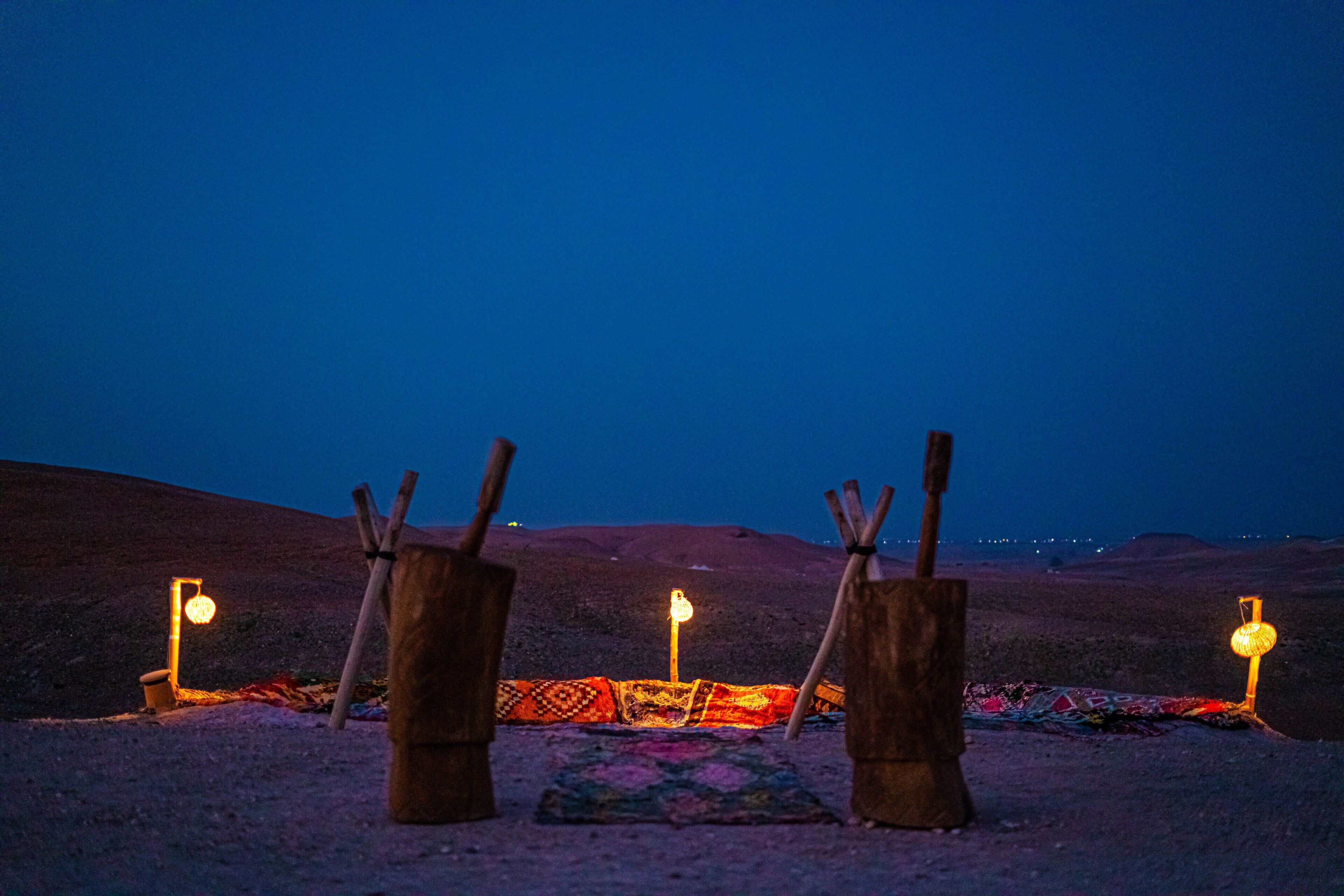 Une scène de désert au crépuscule, avec trois structures en bois et un tapis coloré, le tout illuminé par de petites lumières.