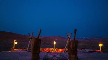 Une scène de désert au crépuscule, avec trois structures en bois et un tapis coloré, le tout illuminé par de petites lumières.